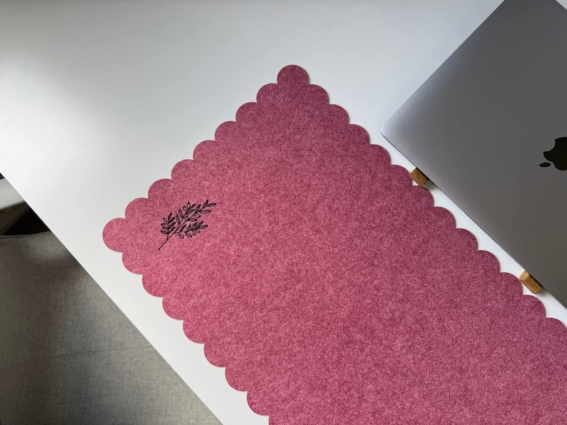 Puede incluir: Una alfombrilla de escritorio rosa polvoriento con un borde festoneado y una ilustraci&oacute;n bot&aacute;nica negra en la esquina. Un ordenador port&aacute;til plateado es parcialmente visible en el lado derecho de la alfombrilla, creando un entorno de trabajo.