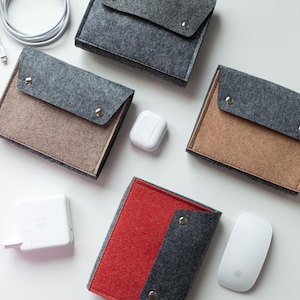 May include: Four rectangular felt pouches with metal snap closures in charcoal, tan, and red colors, accompanied by a white charging cable, a white power adapter, a white wireless earbud case, and a white computer mouse on a plain surface.