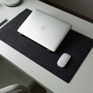 Puede incluir: Una moderna configuración de espacio de trabajo con un portátil plateado, un ratón blanco y una alfombrilla de escritorio de fieltro gris oscuro. El portátil descansa sobre soportes de madera. También se ven un libro y otros suministros de oficina.