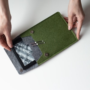 Puede incluir: Una funda de fieltro verde con interior de fieltro gris, que contiene una tableta negra y bolígrafos plateados. La funda está abierta, revelando el contenido. La funda se asegura con dos cierres de presión.