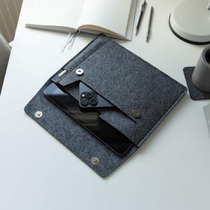 May include: A dark gray felt sleeve case featuring a fold-over flap with two silver-toned magnetic snap closures. The interior compartment holds a black tablet and a black smartphone, with a dedicated side loop for a stylus pen.