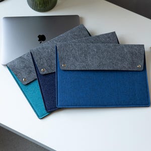 Op de afbeelding: Drie rechthoekige vilten laptophoezen in de kleuren teal, marineblauw en koningsblauw, elk voorzien van een donkergrijze omslagflap die is vastgezet met twee zilverkleurige metalen drukknopen. De hoezen zijn in een waaier vorm gerangschikt.