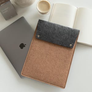 Puede incluir: Una funda para portátil de fieltro en color beige con una solapa gris asegurada con dos broches plateados. Un portátil gris con el logotipo de Apple es visible junto a la funda. Una taza de café y un cuaderno abierto también están en la imagen.