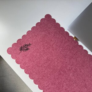 Puede incluir: Alfombrilla de escritorio rosa polvoriento con borde festoneado y dise&ntilde;o bot&aacute;nico negro. Un port&aacute;til plateado es parcialmente visible en el lado derecho, sugiriendo un espacio de trabajo.