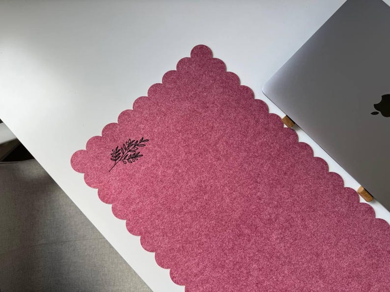 Puede incluir: Alfombrilla de escritorio rosa polvoriento con borde festoneado y dise&ntilde;o bot&aacute;nico negro. Un port&aacute;til plateado es parcialmente visible en el lado derecho, sugiriendo un espacio de trabajo.