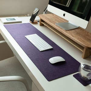 May include: A white desk with a purple desk mat, a white keyboard, a white mouse, a glass of water on a coaster, and a wooden monitor stand.