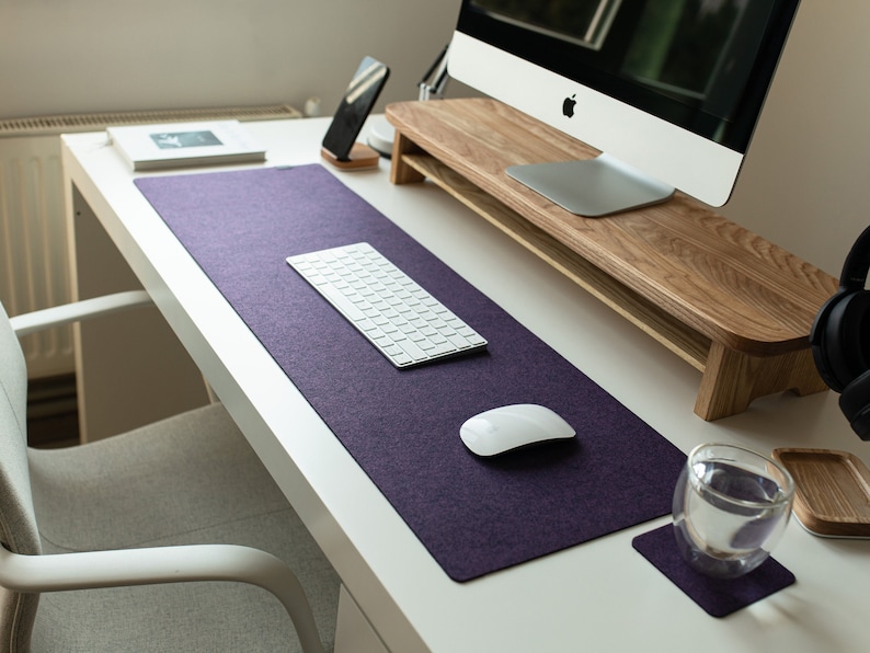 May include: A white desk with a purple desk mat, a white keyboard, a white mouse, a glass of water on a coaster, and a wooden monitor stand.