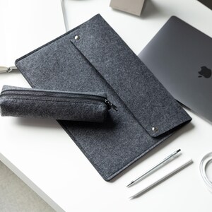 May include: A dark gray felt laptop sleeve with a fold-over flap secured by a silver metal snap button, accompanied by a matching rectangular felt pencil case featuring a black zipper closure, positioned alongside a laptop and two pens.