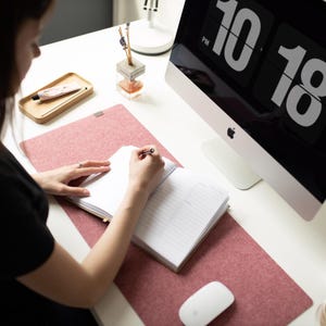 May include: A person is writing in a notebook on a pink desk mat. A computer monitor with a digital clock displaying 10:18 PM is in the background.