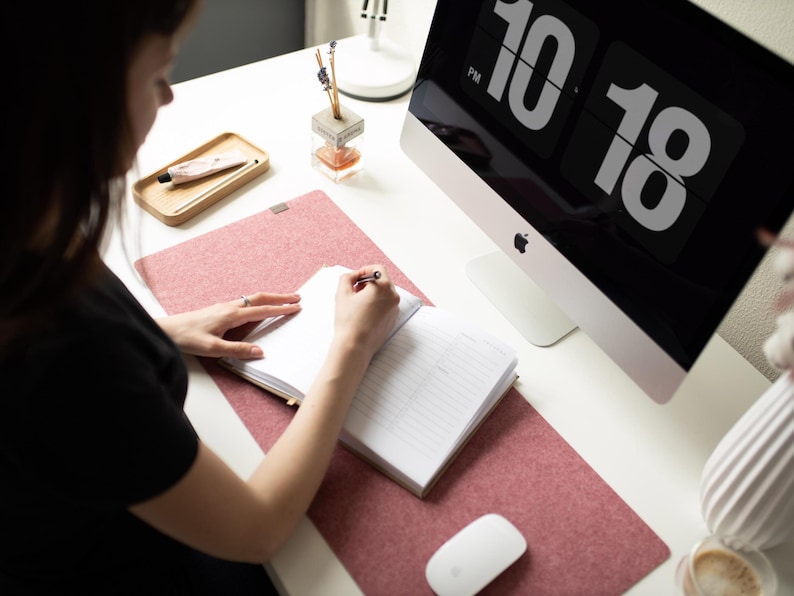 May include: A person is writing in a notebook on a pink desk mat. A computer monitor with a digital clock displaying 10:18 PM is in the background.