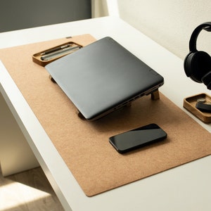 May include: A brown cork desk mat with a laptop, a smartphone, and a headphone stand with black headphones. The stand is made of wood and has a small compartment for storing items.