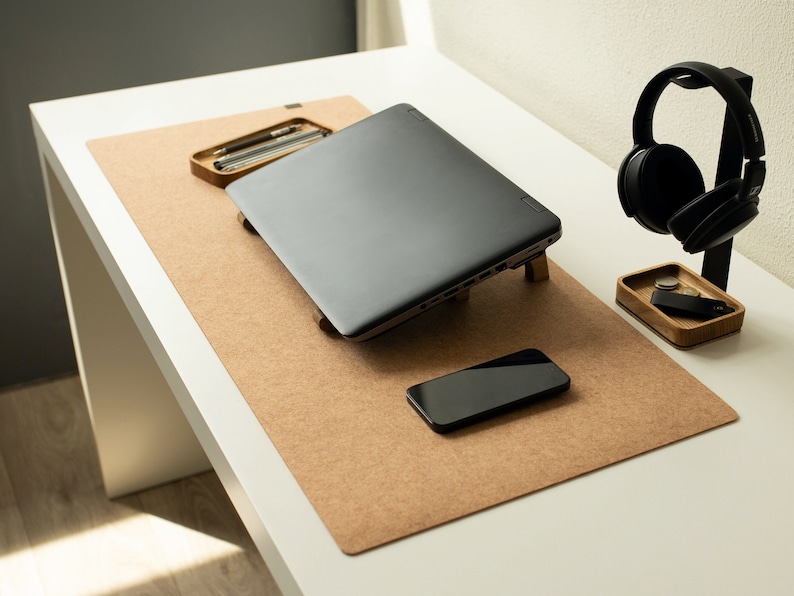 May include: A brown cork desk mat with a laptop, a smartphone, and a headphone stand with black headphones. The stand is made of wood and has a small compartment for storing items.