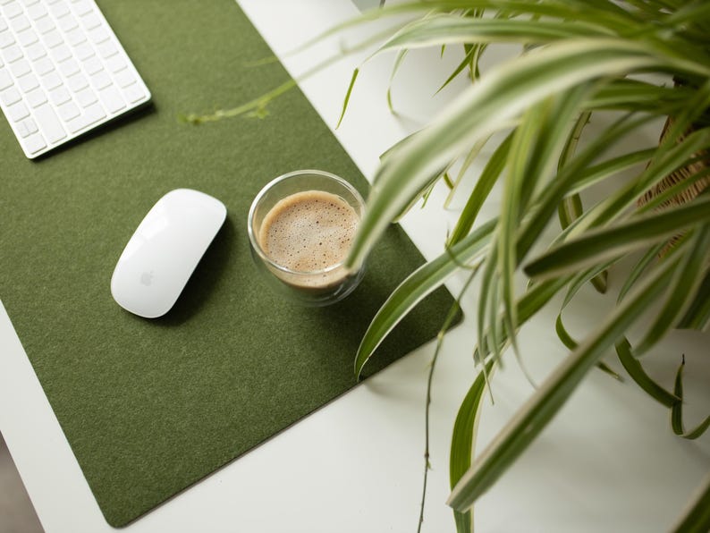 May include: A green felt desk mat with a white computer mouse and a glass of coffee on it. A green plant with white stripes is in the background.