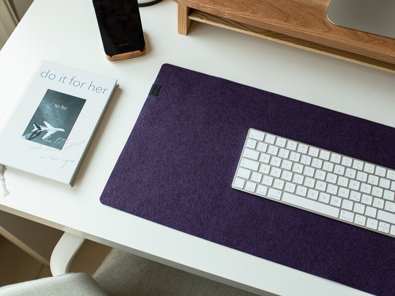 May include: A purple felt desk mat with a white wireless keyboard on top. The mat is on a white desk with a small book that says "do it for her" and a black cell phone on a wooden stand.