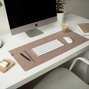May include: A white desk with a brown felt desk mat, a white wireless keyboard, a white wireless mouse, two black pens, a white computer monitor, a white lamp, a white cup, a wooden tray with a white wireless earbud case, and a brown notebook.