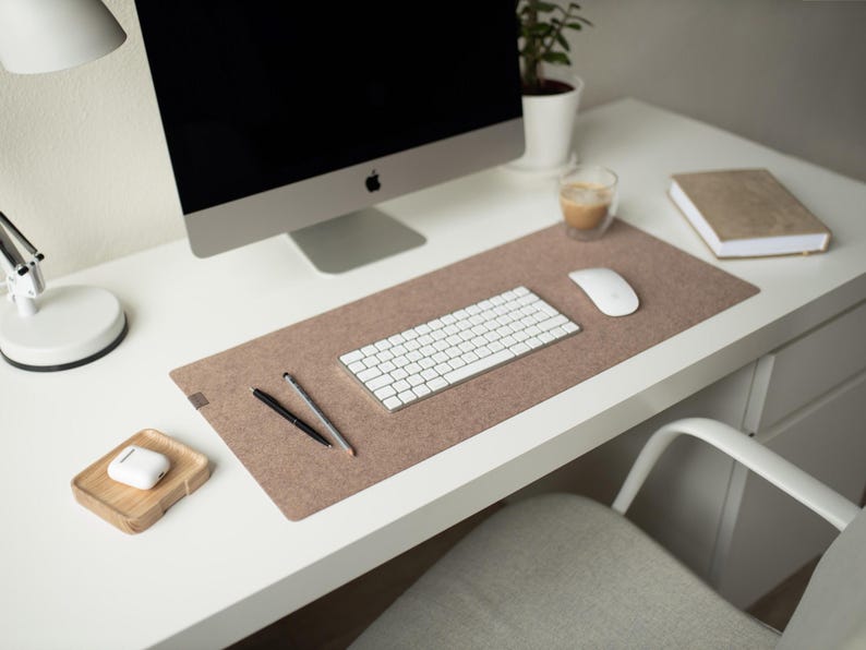 May include: A white desk with a brown felt desk mat, a white wireless keyboard, a white wireless mouse, two black pens, a white computer monitor, a white lamp, a white cup, a wooden tray with a white wireless earbud case, and a brown notebook.