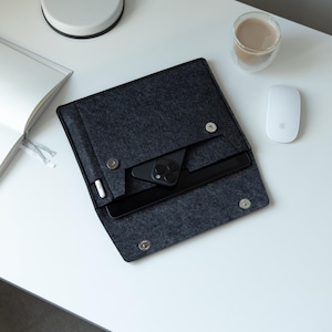 Puede incluir: Una funda de tableta de fieltro gris oscuro con un teléfono negro y un lápiz óptico en el interior, asegurados con cierres de botón. Un cuaderno blanco, una taza de café y un ratón blanco están sobre una superficie blanca.