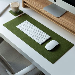 May include: A rectangular olive green felt desk mat with rounded corners. The mat serves as a base for a white wireless keyboard and a white computer mouse. A small matching square felt coaster is positioned to the left of the desk mat.