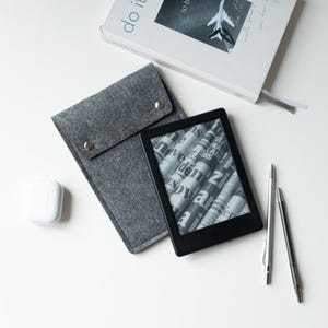 May include: A black e-reader displaying a newspaper graphic, a grey felt sleeve with two silver snap buttons, a white wireless earbud case, two metallic pens, and a white book titled "do it" with an airplane image on the cover.