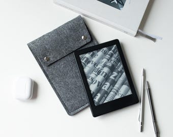 May include: A black e-reader displaying a newspaper graphic, a grey felt sleeve with two silver snap buttons, a white wireless earbud case, two metallic pens, and a white book titled "do it" with an airplane image on the cover.