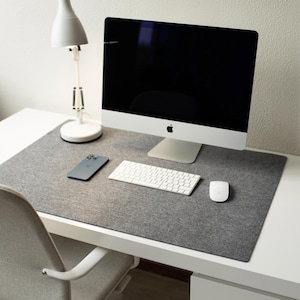 May include: A modern workspace featuring a silver iMac desktop computer, a white keyboard, and a white mouse on a gray felt desk mat. A smartphone and a white desk lamp are also visible. The desk is white, and a gray chair is in the foreground.