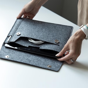 Op de afbeelding: Grijze vilten tablethoes met een zwarte tablet erin. De hoes heeft een vak voor accessoires en een magnetische sluiting. Een witte kabel en stylus zijn zichtbaar. De hoes is open en onthult de binnenkant.
