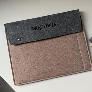 Könnte beinhalten: Eine rechteckige Laptoptasche aus Filz mit einem hellbraunen Hauptteil und einer dunkelgrauen Umschlagklappe. Die Klappe ist mit zwei silberfarbenen Metalldruckknöpfen versehen und zeigt das Wort BRADFORD in einer dunklen Schriftart.