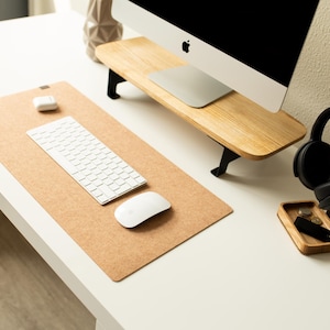 May include: A white computer keyboard and mouse sit on a cork desk mat. A pair of black headphones are on a wooden stand with a small wooden tray. The computer monitor is on a wooden stand.