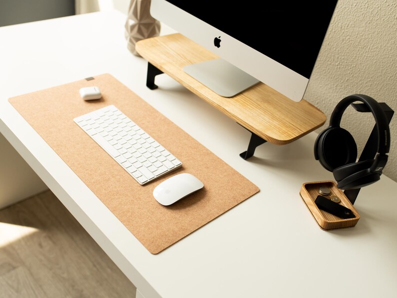 May include: A white computer keyboard and mouse sit on a cork desk mat. A pair of black headphones are on a wooden stand with a small wooden tray. The computer monitor is on a wooden stand.