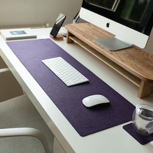 Puede incluir: Una alfombrilla de escritorio rectangular de fieltro morado con esquinas redondeadas, colocada sobre una superficie de escritorio blanca. La alfombrilla sirve de base para un teclado inalámbrico blanco y un ratón de ordenador blanco.