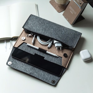 Puede incluir: Organizador tecnológico de fieltro gris con cremallera, que contiene una tableta, un lápiz óptico, cables de carga y un adaptador de carga blanco. Un par de auriculares inalámbricos blancos están cerca. El organizador está abierto, mostrando su contenido.