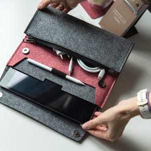 Może przedstawiać: Szare filcowe etui na tablet z czerwonym wnętrzem, czarnym zamkiem błyskawicznym i zatrzaskami. Etui mieści tablet, biały rysik i kable do ładowania. Etui jest otwarte, ukazując zawartość.