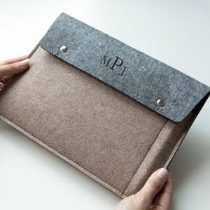 Può includere: Una custodia per laptop in feltro grigio e beige con chiusura a patta. La patta è grigia e fissata con due bottoni a pressione argentati. Le iniziali "MPJ" sono incise sulla patta. La custodia è tenuta da due mani.