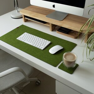 May include: A green felt desk mat with a white keyboard, a white mouse, and a glass cup with a brown beverage on a white desk.
