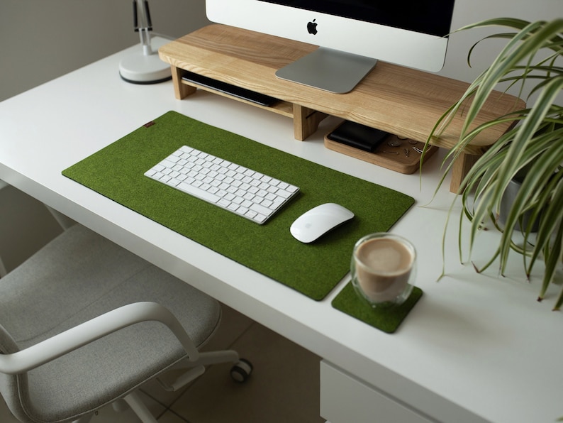 May include: A green felt desk mat with a white keyboard, a white mouse, and a glass cup with a brown beverage on a white desk.