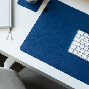 Puede incluir: Una alfombrilla de escritorio de fieltro azul con un teclado blanco sobre un escritorio blanco. También hay un posavasos a juego y un cuaderno en el escritorio. La alfombrilla de escritorio es rectangular y tiene una superficie texturizada.