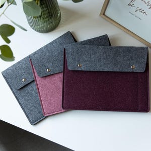 May include: Three rectangular felt document sleeves with fold-over flaps secured by silver metal snaps. The sleeves are colored in dark gray, dusty rose, and deep burgundy, each featuring a contrasting dark gray felt flap and a structured design.