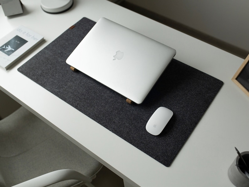 May include: A gray felt desk mat with a silver laptop computer and a white wireless mouse. The laptop has a silver Apple logo on the lid. The desk mat is on a white desk.