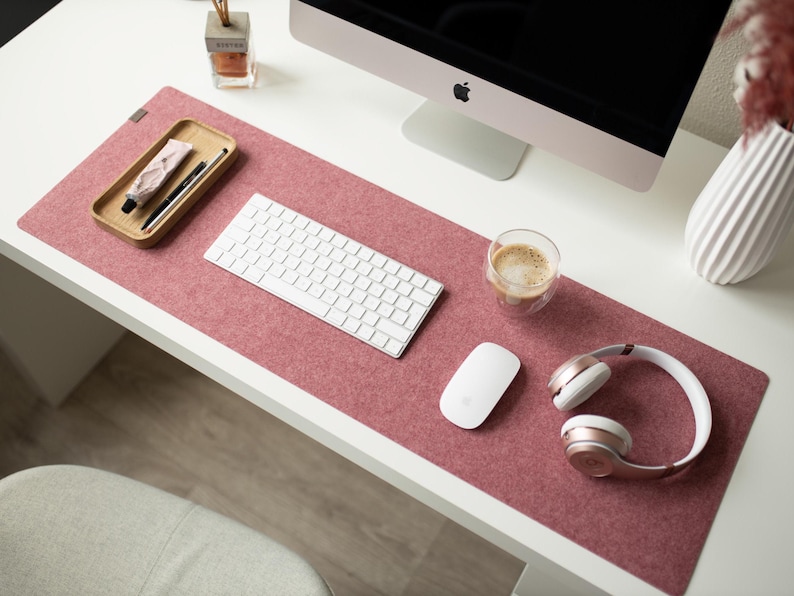 May include: A pink felt desk mat with a white keyboard, a white mouse, a glass of coffee, and rose gold headphones.