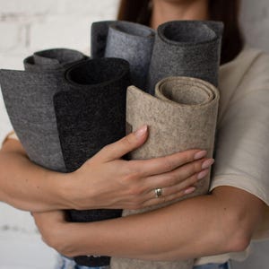 May include: A collection of rolled felt mats in various shades of gray, charcoal, and beige. The mats have a thick, textured surface and are held together in a bundle. Each mat is cylindrical in shape, showcasing a dense, fibrous material.