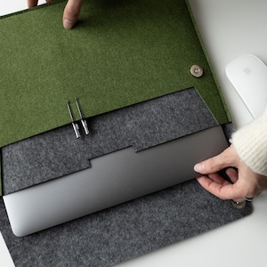 Puede incluir: Una funda para portátil de fieltro verde con interior gris. La funda está abierta, revelando un portátil plateado. Un ratón blanco y un cable de carga son visibles sobre la superficie blanca.
