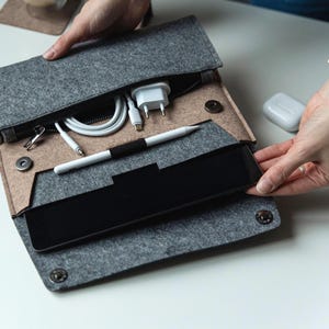 Könnte beinhalten: Eine graue Filz-Tablettasche mit einem Reißverschlussfach, das ein weißes Ladegerät, ein Kabel und einen Stift enthält. Die Tasche ist offen und zeigt ein schwarzes Tablet. Die Tasche wird mit Druckknöpfen verschlossen.