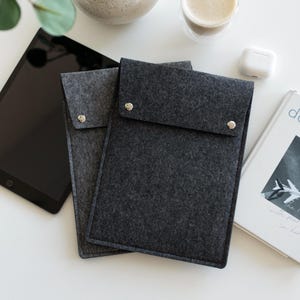 May include: Two rectangular felt sleeves for electronic devices, one in light gray and one in dark charcoal gray. Each sleeve features a fold-over flap secured with a single silver-toned metal snap button closure at the center of the flap edge.