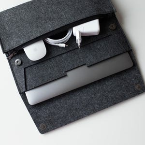 May include: A gray felt laptop sleeve with a zipper closure and a built-in pocket for accessories. The sleeve is open and a laptop, a white wireless mouse, and a white power adapter are visible inside.