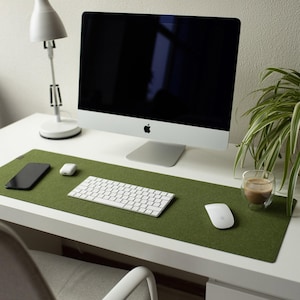 May include: A rectangular olive green felt desk mat positioned on a white desk surface. The mat serves as a base for a white wireless keyboard, a white computer mouse, a black smartphone, and a small white charging case for wireless earbuds.