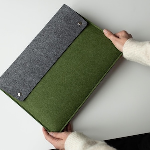 Puede incluir: Una funda rectangular para portátil hecha de fieltro verde oliva con una solapa de fieltro gris oscuro. La solapa se asegura con dos botones de presión de metal plateado. La funda presenta un diseño estructurado con bordes definidos.