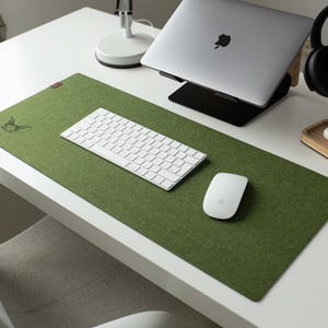 May include: A green felt desk mat with a white keyboard, a white mouse, and a small wooden stand with a white wireless charging pad. The desk mat has a small embroidered design in the top left corner.