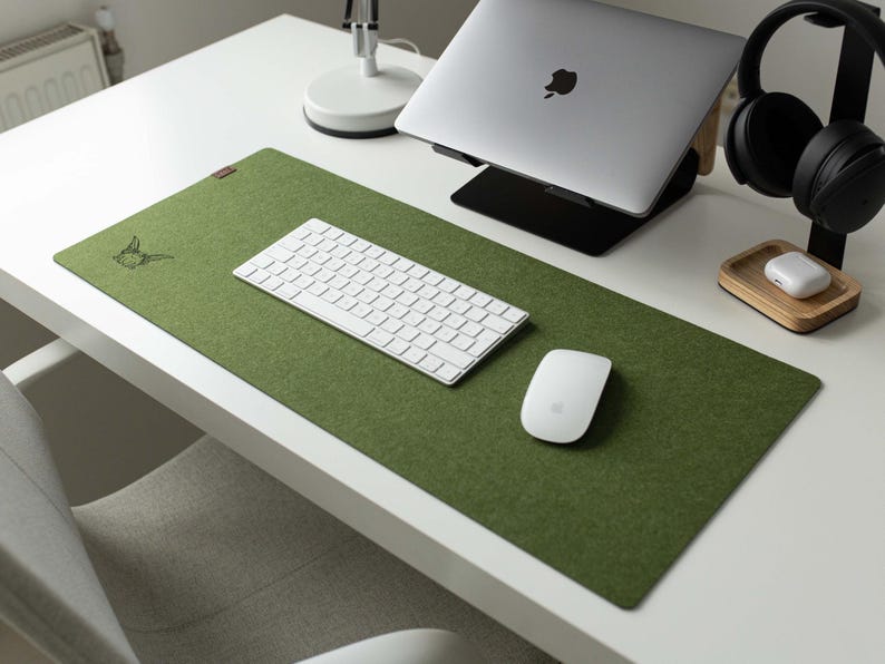 May include: A green felt desk mat with a white keyboard, a white mouse, and a small wooden stand with a white wireless charging pad. The desk mat has a small embroidered design in the top left corner.