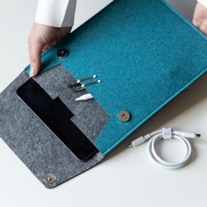 Könnte beinhalten: Türkis-graue Laptop-Hülle aus Filz mit Magnetverschluss. Die Hülle ist geöffnet und zeigt ein Tablet, Stifte und einen weißen Stift. Ein weißes Ladekabel und eine Computermaus sind ebenfalls sichtbar.