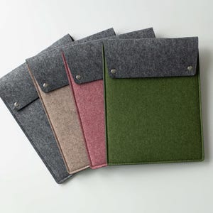 Puede incluir: Fundas para portátiles de fieltro en gris, beige, rosa y verde. Cada funda tiene una solapa gris asegurada con broches plateados. Las fundas están dispuestas en abanico, mostrando sus colores. Medidas en centímetros.